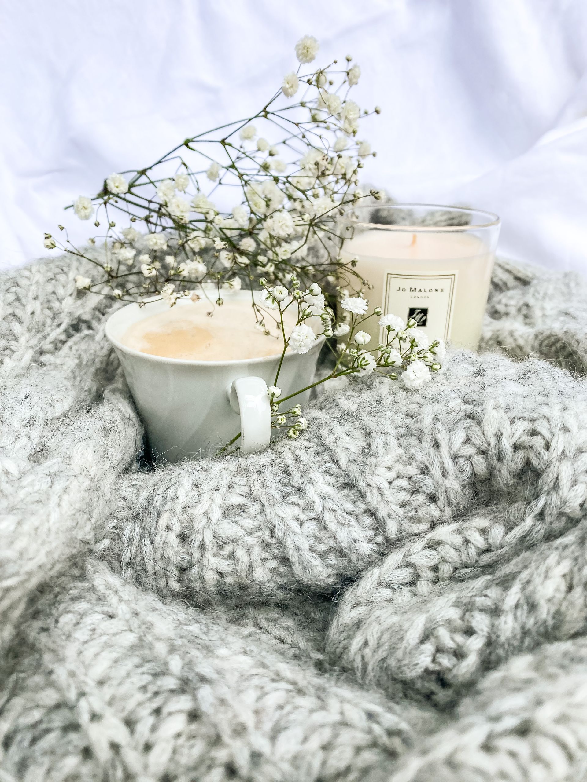 A warm and cozy setting with a coffee cup, candle, flowers, and a knit blanket.